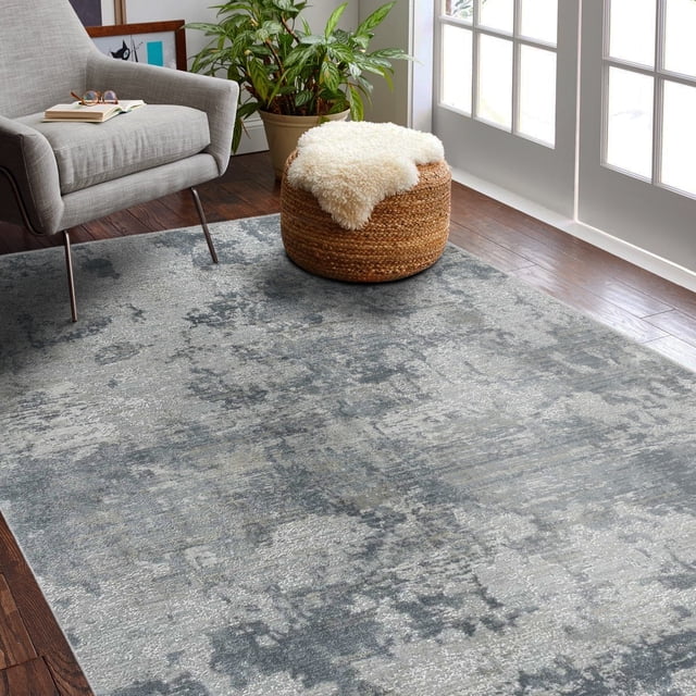 Rug Branch Melo Collection Modern Abstract Scandinavian Indoor Area Rug with a rectangular shape and muted gray-blue abstract pattern, placed on dark hardwood floor in a living room. The rug is under a gray armchair and a woven ottoman with a white sheepskin throw, near a green potted plant and glass doors. Suitable for living rooms and hallways as a durable indoor floor covering.