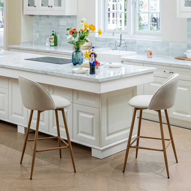 Homy Casa set of 2 modern bar stools with beige fabric upholstery, 26-inch seat height, curved backrests with cross-stitch detailing, wooden legs with footrests, and 360-degree swivel seats, placed at a white marble kitchen island in a bright kitchen with flowers and kitchenware, also suitable for casual workspace seating.