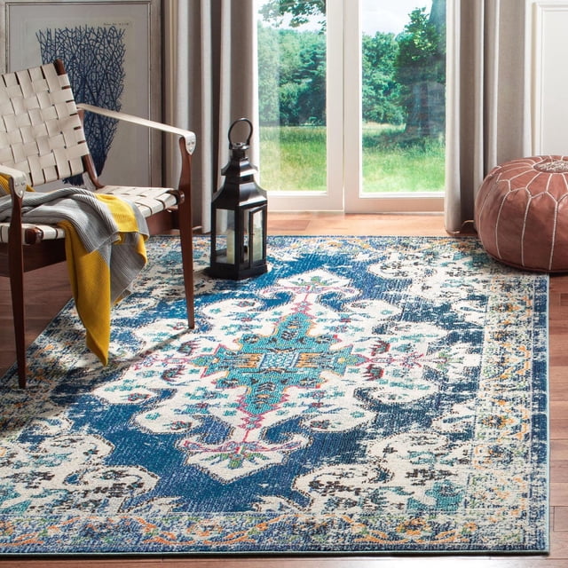 Safavieh Madison Richmal Vintage Traditional Area Rug with a blue and cream medallion pattern and vintage distressed look, power loomed for durability, placed on hardwood flooring in a bright room with beige curtains, a woven strap chair with a yellow and gray throw, a black lantern, and a brown leather pouf near glass doors showing green trees, ideal for classic and transitional home decor.