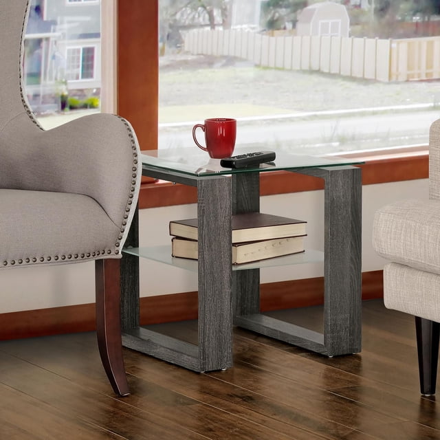 Caleb Grey Wood Side End Table with Glass Shelf featuring a grey oak finish wood-look frame, clear tempered glass top, and frosted glass lower shelf holding two books, placed between a beige armchair and sofa on a hardwood floor near a window, ideal for displaying decor or storing reading materials.