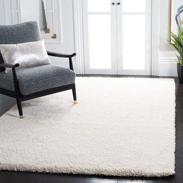 Safavieh Milan Harlow Tapis Shag Uni en Ivoire Éclatant, Tissé Mécaniquement avec des Fils Synthétiques Durables, Posé sur des Planchers de Bois Franc Sombres à Côté d'Un Fauteuil en Bois Noir avec Rembourrage à Motifs et un Coussin Lombaire Argenté, Ajoutant une texture Douce et un style Européen Contemporain à la Déco de la Maison.