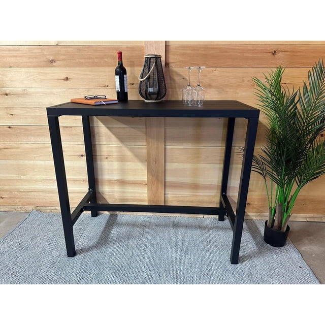 Highland bar table 20''x43'' with a durable black aluminum top and sturdy metal frame, set against a light wood paneled wall. The table holds a bottle of red wine, a black lantern with rope handle, two inverted wine glasses, a closed notebook, and reading glasses. It stands on a gray woven rug next to a potted green plant, suitable as a compact bar table, dining spot, or console table for small spaces.