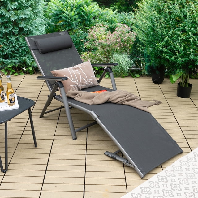 Costway Patio Folding Aluminum Lounge Chair Chaise with adjustable backrest and armrests, designed for outdoor use. The grey aluminum frame supports a black quick-drying fabric seat, and it includes a removable headrest pillow for added comfort. Ideal for patios, poolside, or backyards, the chair is lightweight and foldable for easy storage. Shown on a wooden patio surrounded by greenery, with a side table holding beverages nearby.