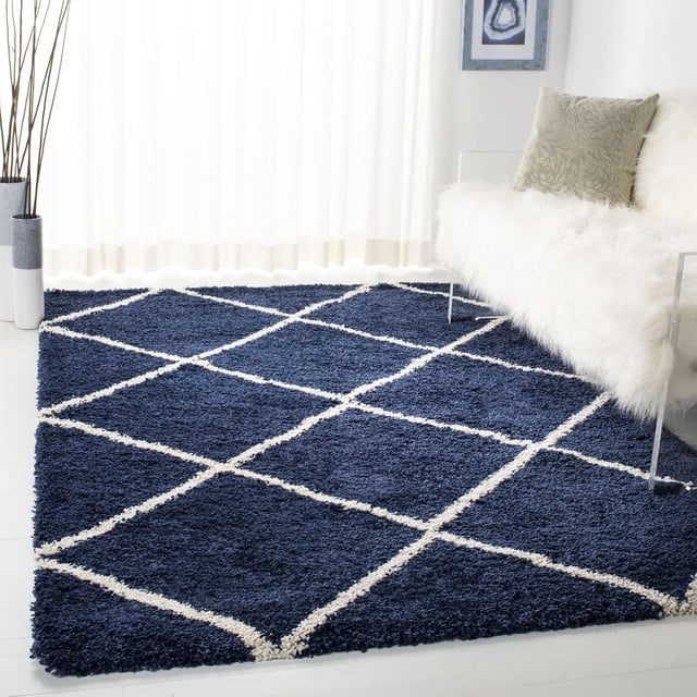 Safavieh Hudson Amias Geometric Shag Area Rug with a thick navy blue shag pile and ivory geometric tile pattern, placed on a light floor beside a white faux fur accent chair with a beige pillow, near sheer curtains in a modern living space. Ideal for adding texture and comfort to urban or contemporary décor.