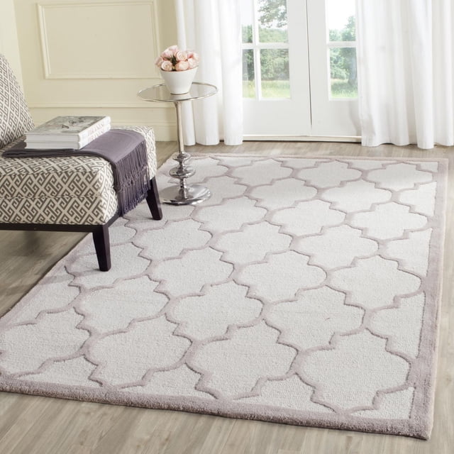 Safavieh Cambridge Kevin Geometric Area Rug in a light neutral colour with a raised interlocking geometric pattern, hand-tufted wool texture, placed on light hardwood flooring in a bright room with white curtains and a patterned accent chair with a throw and books.