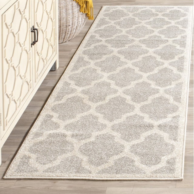 Safavieh Amherst Roderick Geometric Area Rug with a light beige and cream quatrefoil pattern, power-loomed polypropylene construction with cut pile texture, ideal for high-traffic indoor or outdoor spaces, displayed on a light wood floor next to a cream geometric cabinet and woven basket with a yellow throw.