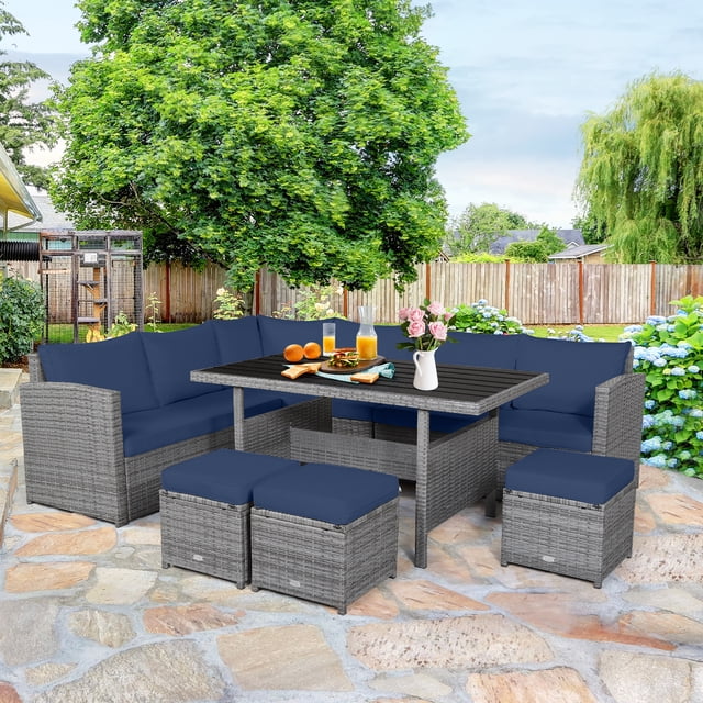 Patiojoy Ensemble de Conversation en Osier d'Extérieur, 7 Pcs, avec Coussins Bleu Marine Exposés dans un Décor de Cour Arrière. Cet Ensemble de Mobilier de patio Comprend Trois Chaises d'Angle, Trois ottomans et une table Rectangulaire avec un Dessus en Bois Plastique. le Cadre en Rotin Gris Mélangé Complète les Coussins Bleu Marine, Créant un Ensemble de Salle à Manger d'Extérieur Moderne et Invitant. la table Est Conçue avec des Aliments et des Boissons, Entourée d'Un Feuillage Luxuriant et d'Une Clôture en Bois.