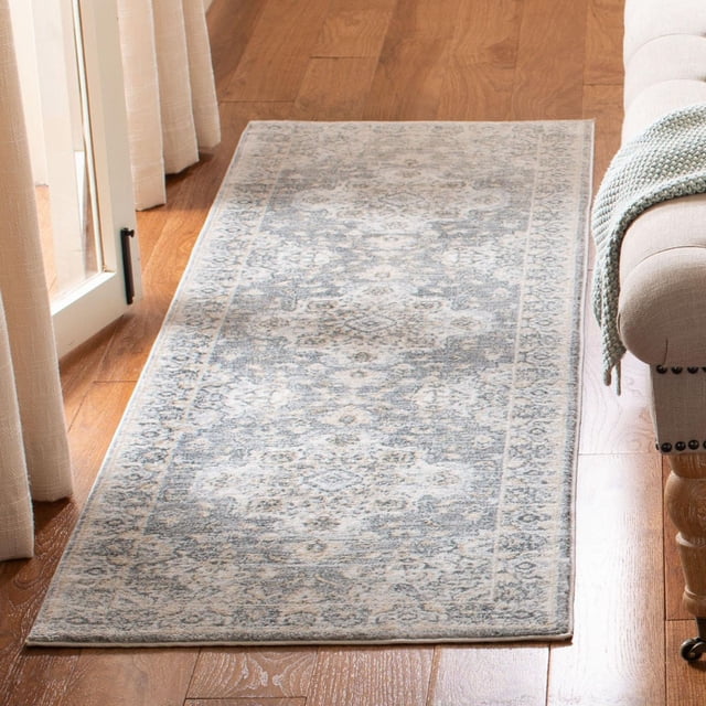 SAFAVIEH Isabella Tamzen Floral Bordered Area Rug in soft grey and blue floral medallion pattern with ornate border, power-loomed synthetic yarn and medium cut pile, on hardwood floor beside beige sofa with knitted throw, ideal for traditional or classy-casual decor and suitable as a decorative hallway runner.