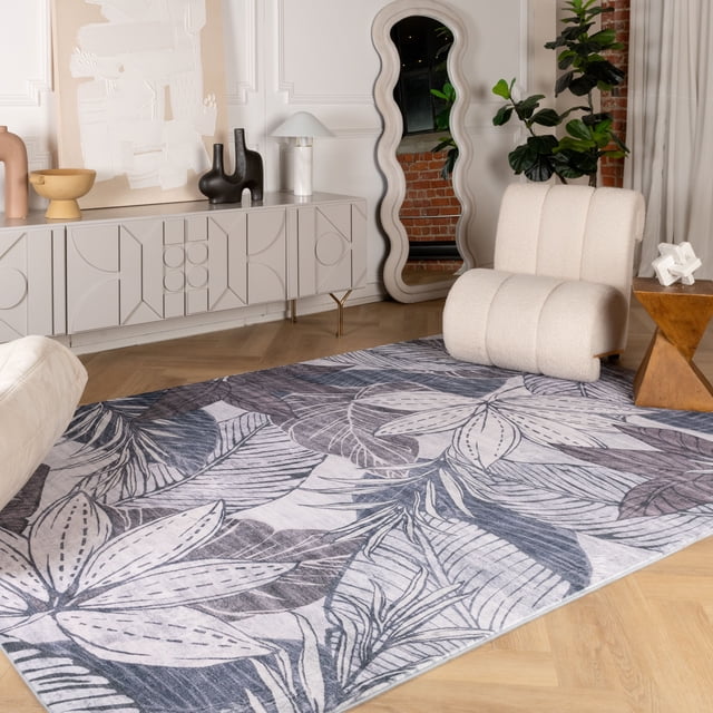 Rug Branch Modern Floral Indoor Area Rug in grey with large floral and leaf patterns in shades of grey and white, placed on light hardwood flooring in a modern living room with a cream boucle lounge chair, wooden geometric side table, wavy full-length mirror, and white decorative sideboard with vases and lamp, ideal for enhancing interior decor with a soft, plush texture.