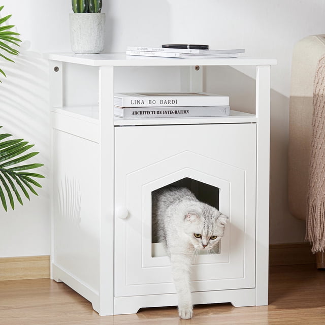 Cat Litter Box Enclosure by LIVINGbasics, a white decorative nightstand that doubles as a hidden cat house and indoor pet crate. Features a front-facing arched entry door for cats, a top surface for decor or storage, and a handy shelf for organizing items. Made of solid wood, MDF, and metal hinges, it measures 19.5 inches wide, 21.25 inches deep, and 25.18 inches high. A grey cat is seen exiting the enclosure, which is placed in a modern indoor setting with books and a plant on top.