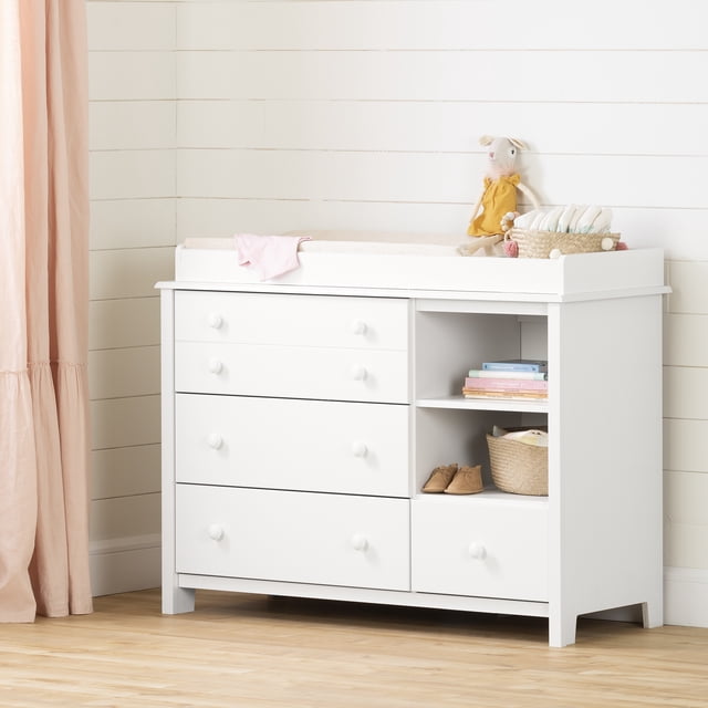 South Shore Little Smileys table à Langer avec Rebord Amovible en Blanc Pur, avec Quatre Tiroirs avec Poignées à Bouton en Bois, Deux Étagères de Rangement Ouvertes avec une Tablette Détachable et un Rebord Surélevé pour des Changements de Couches Sécuritaires, Style avec un Matelas à Langer beige, un Tissu Rose, un Toutou en Robe Jaune, un Panier de Couches, des Chaussures pour Bébé et des Livres pour Enfants, Posé contre un Mur shiplap Blanc et un Plancher de Bois Clair.