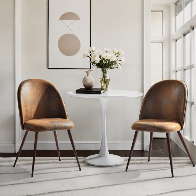 Homy Casa 2-Pack Armless Upholstered Accent Chairs with Round Back in brown faux suede, shown front-facing beside a white round pedestal table with a vase of white flowers and a beige decorative vase on a book, set in a bright living room seating area with large windows and minimalist wall art, suitable for compact spaces.