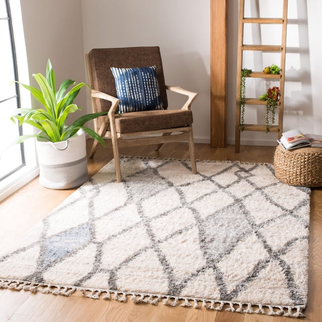 SAFAVIEH Berber Fringe Hattie Tapis Shag Géométrique avec pile de 1,25 Po en Motifs Géométriques Tribaux Crème et Gris et Bords à Franges, Posé sur un Plancher de Bois Franc Clair. le Tapis Est Présenté dans une Pièce Lumineuse avec un Fauteuil en Bois Brun Présentant un Coussin à Motif Bleu, un Panier Tressé avec un Livre Ouvert, une Étagère Échelle en Bois avec des Succulentes, et un Cache-Pot Tressé Blanc et Gris Tenant une Plante Verte. Il Convient Aussi pour Ajouter une Touche Bohème-Chic Douillette aux Espaces de Vie.