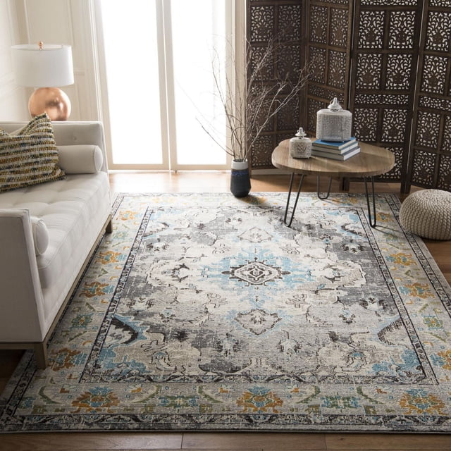 Safavieh Monaco Toria Traditional Area Rug in a bright living room with a beige tufted sofa and multicolored pillow. The rug features a central medallion in blue, grey, and cream with floral and geometric borders in yellow and green. A round wooden coffee table with black metal legs holds decorative jars and books, set against a dark carved wooden screen and a tall vase with bare branches on light hardwood floors. Its durable polypropylene weave is suitable for both country and city homes and often used as a stylish accent in living spaces.