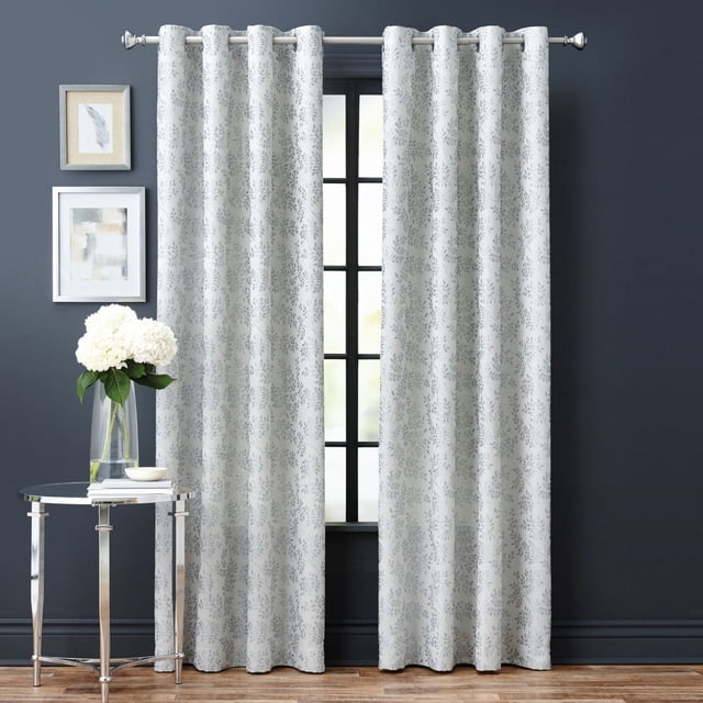 Hometrends Everly 84" light filtering grommet window panel in grey with a leaf-inspired textured fabric, hanging on a silver curtain rod over a black-framed window in a room with dark grey walls, hardwood floor, glass side table with white flowers, and framed artwork, ideal for softening sunlight and enhancing decor.