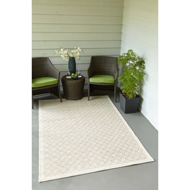 ECARPET Eve Contemporary Area Rug with a tan and ivory geometric basketweave pattern made from stain-resistant polypropylene fibers, placed on a grey concrete floor in a porch setting with two brown wicker chairs with green cushions, a matching wicker side table holding a blue vase with white flowers and a bowl of green apples, and a potted green plant in a black planter against white horizontal siding walls, suitable for indoor and outdoor use.