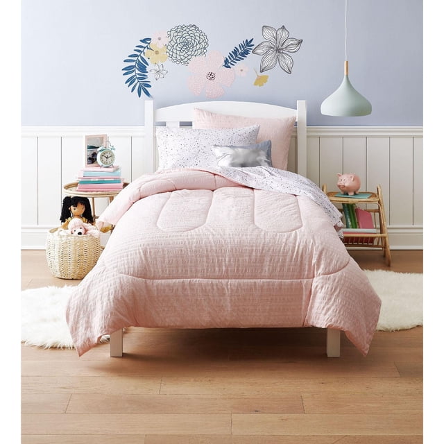 Mainstays Kids Sersuckr Bed in a Bag with a twin bed featuring a soft pink microfiber comforter, reversible white fitted sheet with black speckles, and matching pillowcases, set in a child's room with floral wall decals, wicker basket with dolls, and light wood flooring, machine washable and suitable for all seasons.
