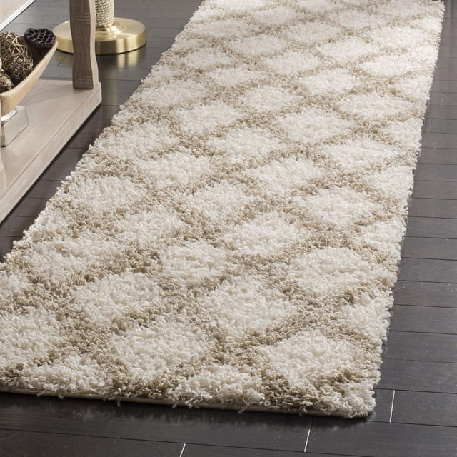 Safavieh Layla Geometric Plush Shag Area Rug with a beige and cream diamond pattern, dense synthetic fiber pile on dark hardwood floor beside a light wood console table and brass lamp base, ideal for high-traffic areas and adding texture to modern home decor.