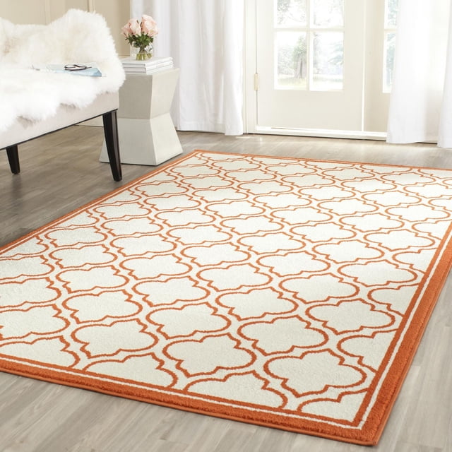 Safavieh Amherst Wilson Geometric Area Rug with a cream base and orange geometric trellis pattern, power loomed polypropylene construction for durable indoor-outdoor use, shown on light wood flooring in a bright room with white curtains and a modern chair.