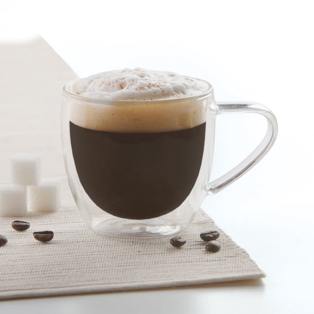 Cappuccino Mug Double Wall made of clear borosilicate glass with double-wall insulation holding dark coffee topped with frothy foam, placed on a beige placemat with coffee beans and sugar cubes, designed for thermal shock resistance and suitable for home or café use.