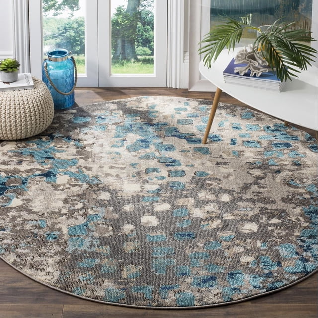 Safavieh Monaco Driskoll Abstract Area Rug, a round polypropylene accent rug with durable blue, beige, and gray abstract patterns, placed on a wooden floor near glass doors with natural light, styled with a white mid-century modern table holding a plant and books, and a knitted pouf with a blue lantern, ideal for adding Bohemian-chic flair to living spaces.