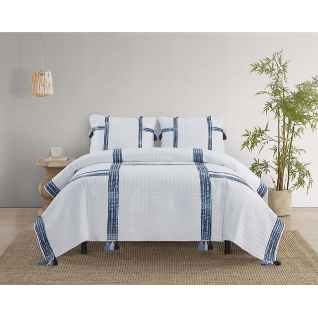 Luana 3 Piece Quilt Set with white quilt and matching shams featuring blue shibori stripe appliqué and grid pattern stitching, edged with multicolor blue tassels, displayed on a bed in a neutral bedroom with wooden side table, hanging wicker lamp, and potted bamboo plant, ideal for refreshing bedroom décor.