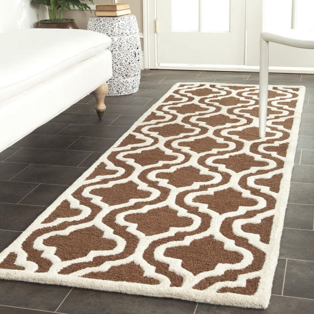 Safavieh Cambridge Kirsten Geometric Area Rug rectangular runner with brown and white geometric pattern, hand-tufted wool featuring plush cut and high/low loop pile texture, placed on dark tile flooring in a bright room with white furniture and ceramic stool, ideal for hallways or entryways.