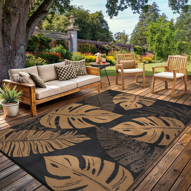 Garvee 8'x10' Outdoor Rug Waterproof Plastic Straw Rug with a modern tropical leaf design in black and brown, ideal for patios, RVs, and beach outings. The UV-resistant and reversible rug is displayed on a wooden deck surrounded by outdoor furniture and vibrant greenery.