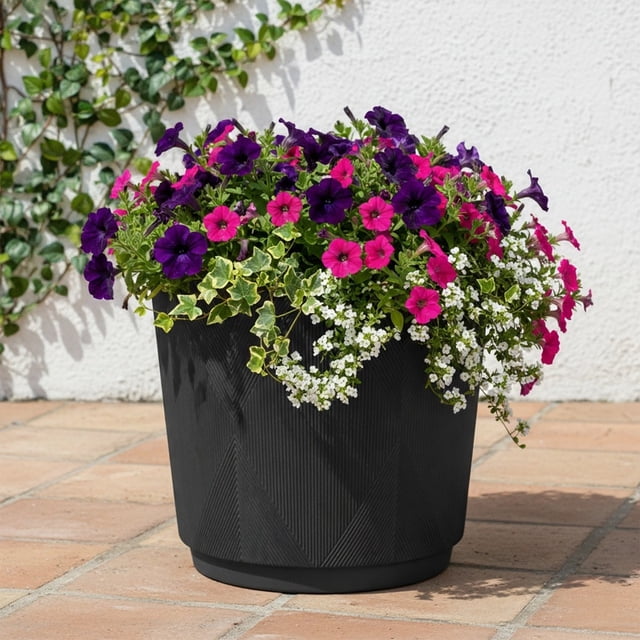 Better Homes & Gardens 16in Cappriccio Resin Planter in black with a geometric textured finish, lightweight and weather-resistant, holding purple and pink petunias, white alyssum, and trailing variegated ivy on a tiled patio outdoors, suitable for indoor or outdoor gardening.
