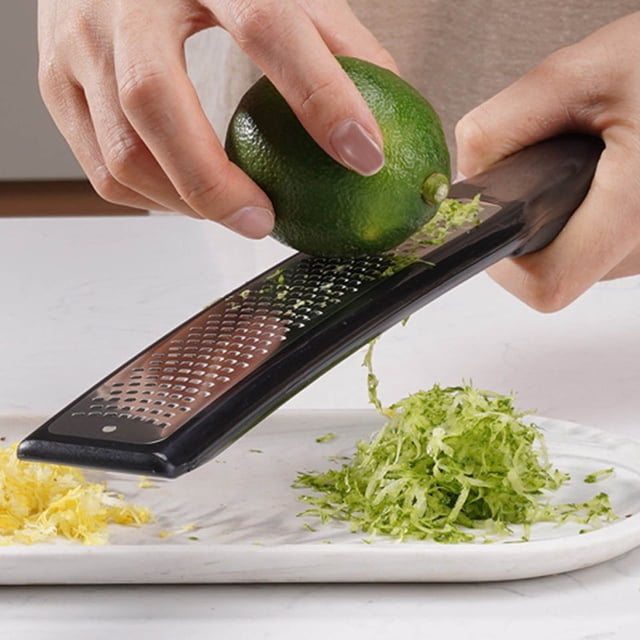 Czzoypi Râpe à Fromage (D), une Râpe Polyvalente en Acier Inoxydable avec une Poignée Ergonomique Noire, Montrée Râpant une lime. le Zeste de lime Râpé Est Récolté sur une Assiette Blanche à Côté du Zeste de Citron. la Râpe Présente des Lames Tranchantes et Résistantes à la Rouille et Est Livrée avec un Couvercle Protecteur et une Brosse de Nettoyage pour un Entretien Facile.