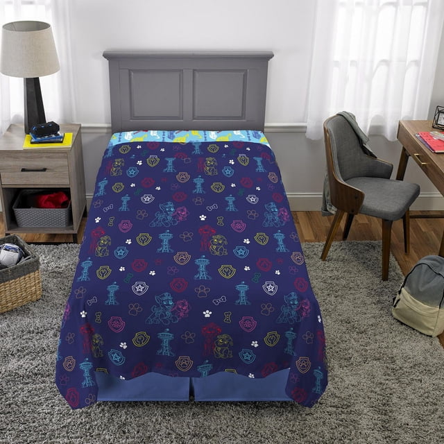 Pat' Patrouille Ensemble de Draps Simple « So Fun » avec Draps-Housses et Draps Plats Bleu Marine Présentant des Contours Colorés des Personnages Pat' Patrouille Chase, Rocky, Zuma, Rubble et Marshall, Ainsi Que des Empreintes de Pattes, des Os et des badges, sur un Lit Simple dans une Chambre d'Enfant avec Tête de Lit Grise, Table de Nuit en Bois et Chaise Grise, Convient à la Literie pour Enfants.