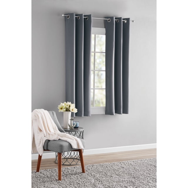 MAINSTAYS Blackout Energy Efficient Grommet Window Curtains Panel in dark grey hanging on a silver rod above a window in a room with light grey walls, a grey chair with wooden legs covered by a white throw, a round metal side table with a white vase of flowers and a blue candle, and a light grey rug on a wooden floor, suitable for blocking light and improving energy efficiency.