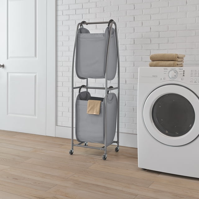 MAINSTAYS Rolling Vertical Laundry Sorter with a sturdy metal frame and two collapsible gray fabric totes featuring ergonomic foam handles, mounted on four caster wheels for easy mobility, placed in a laundry room next to a white front-loading dryer with folded beige towels on top, suitable for maximizing vertical space in compact laundry areas.