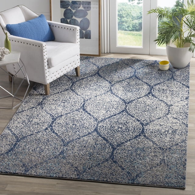 Safavieh Madison Leighton Geometric Area Rug with a blue and cream trellis pattern, power loomed for durability, shown on light wood flooring in a bright room featuring a white nailhead-trimmed armchair with blue cushion, a metal side table, framed abstract art, and a large potted plant by glass doors overlooking greenery, ideal for enhancing contemporary and transitional home decor.