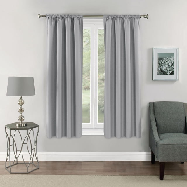 Eclipse Samara 63" Rod Pocket Blackout Panel in light grey with textured tone-on-tone design, hanging on a silver rod over a window. The foam-backed thermal curtain blocks over 99% of light, reduces noise, and saves energy, shown in a living room with grey armchair, metal side table, and lamp. Also suitable for creating a darkened space for media rooms or bedrooms.