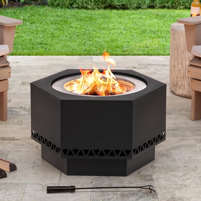 Sunjoy 26 inch Hexagonal Low Smoke Firepit with black hexagonal steel body and triangular ventilation cutouts near the base, featuring a stainless steel combustion chamber with burning wood flames, set on a stone patio between wooden chairs and a side table, including a black fire poker tool in front for outdoor heating.