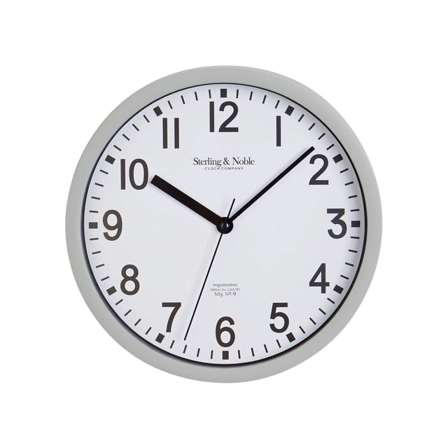 HOMETRENDS Basic Indoor 8.8-inch Gray Analog Round Wall Clock. Front-facing view of an 8.8-inch (22.4 cm) gray plastic framed analog wall clock with glass lens, white inlay, black numerals and white accents; quartz movement; requires 1 AA battery; suitable for kitchens or offices.