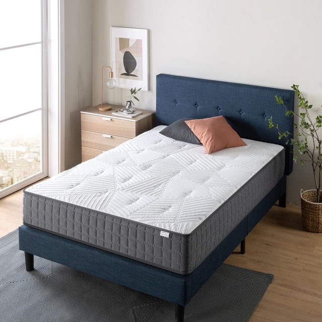 Zinus Spa Sensations 10 inch Charcoal and Copper Hybrid Memory Foam and Spring Mattress on a blue fabric bed frame with tufted headboard in a bright bedroom. The mattress features a white quilted top with geometric stitching and charcoal grey sides. Two pillows, grey and rust-coloured, rest on the mattress. Nearby is a wooden nightstand with a copper lamp, abstract wall art, and a green potted plant by a large window. The mattress includes iCoil technology for motion reduction and cooling graphite and copper gel foam for temperature regulation, suitable for maintaining comfort and freshness.