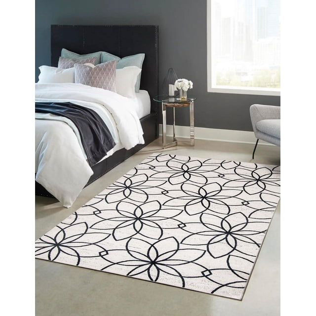 ECARPET Washable Floral Area Rug & Runner for Bedroom, Living Room, Kids Room, Hallway with a low-pile polyester surface featuring a black floral geometric pattern on white, placed in a modern bedroom with a black upholstered bed, blue and patterned pillows, a glass side table with white flowers, and a grey armchair near a large window, ideal for high-traffic indoor spaces.