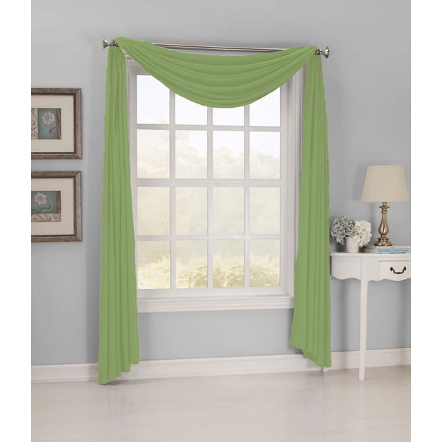 918 Hampton Microfiber Scarf in willow green draped over a silver curtain rod above a white-framed window with multiple panes in a light gray room. The polyester fabric has a smooth texture and rod pocket design, measuring 54 inches wide by 216 inches long. The scarf is machine washable and adds decorative elegance to window treatments and room darkening curtains. The setting includes a white side table with a brass lamp and floral arrangement, plus framed botanical prints on the wall. This scarf is also suitable for layering with other window coverings.