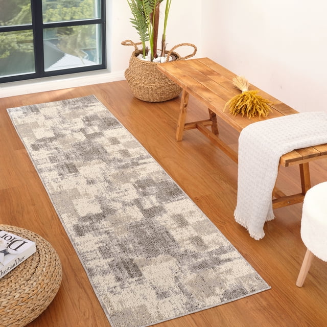 Rug Branch Jules Abstract Indoor Area Rug in grey beige with a modern abstract pattern, laid on a hardwood floor in a bright room. The polypropylene rug features a 0.3-inch pile height and jute backing, placed next to a wooden bench with a light grey throw and dried yellow flowers, near a woven basket planter and a round woven ottoman with a book.