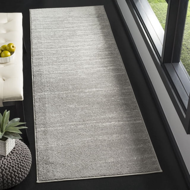 SAFAVIEH Adirondack Esmond Abstract Faded Area Rug, a rectangular accent rug with a subtle abstract faded pattern in light grey tones, made of easy-care polypropylene yarns, placed on a dark floor near a window and a white tufted bench with green apples.