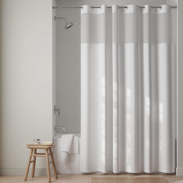 Zenna Home InstaCurtain Fabric Hotel Shower Curtain in white with built-in rustproof grommets and hooks, featuring a sheer top window panel, hanging on a metal rod in a bathroom with light grey herringbone tile and a wooden stool beside the tub, suitable for easy installation without separate hooks.