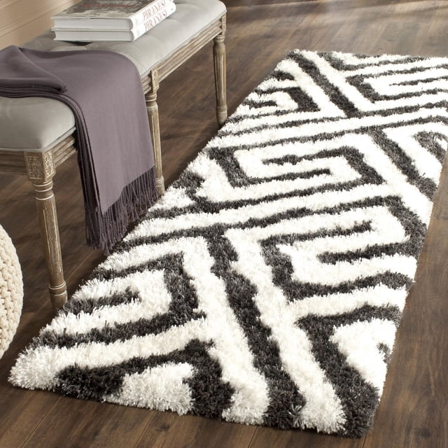 Safavieh Barcelona Dortha Geometric Shag Area Rug featuring black and white Moroccan quatrefoil tile motifs and layered line patterns on a plush shag pile, placed on dark hardwood flooring beside a wooden bench with a gray throw and books, suitable for modern accent décor.