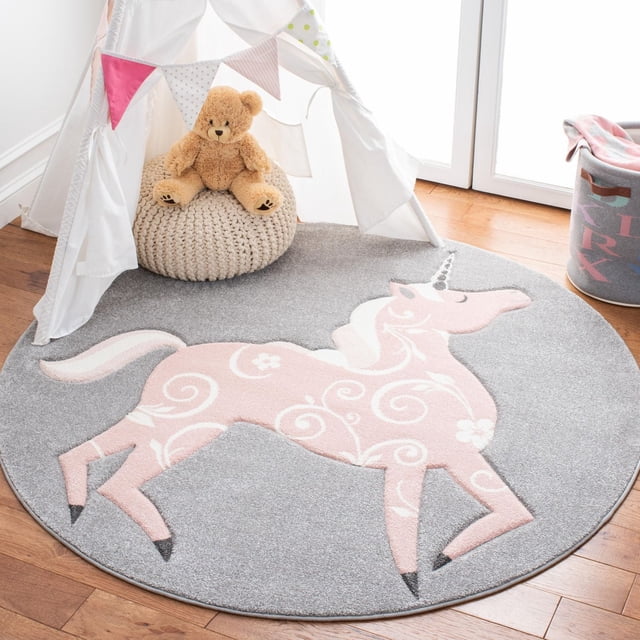 Safavieh Carousel Kids Dalach Unicorn Area Rug with a round grey background and a pink unicorn decorated with white floral swirls, power-loomed for durability, displayed in a child's room setting with a white play tent, beige knitted pouf, teddy bear, and pastel storage bin, suitable for soft, colourful kids' decor.