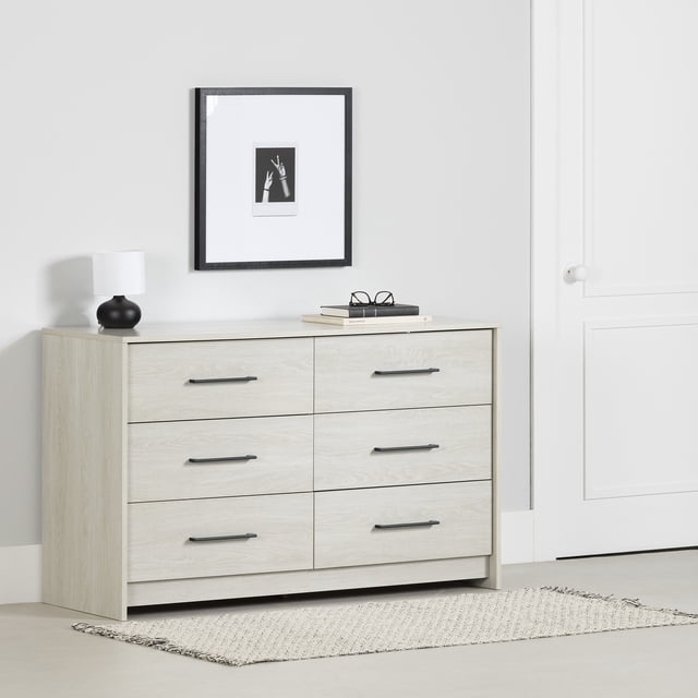 South Shore Londen 6-Drawer Dresser for Adult in winter oak finish with six spacious drawers featuring long black metal handles and metal slides, shown in a minimalist bedroom setting with a black lamp, stacked books, and framed black-and-white hand photo above, placed on a light woven rug, ideal for organized clothing storage.