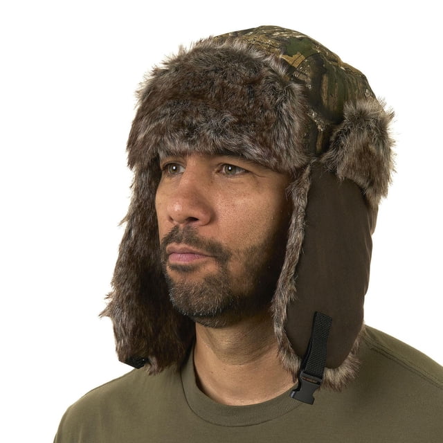 Realtree APX Trapper Hat RT/Brown shown in a 3/4 profile view with camo polyester outer fabric and brown faux fur trim on ear flaps and front. It includes a quick-release buckle chin strap and sherpa fleece lining for warmth, suitable for men's hunting clothing to protect against cold and wind.