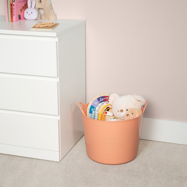 Neatfreak Kids Large Flexible Bin w/ Handles in peach flexible plastic with built-in handles, placed on beige carpet beside white dresser, containing children's toys including two teddy bears and colorful educational clocks, suitable for lightweight storage and easy transport in kids' rooms.