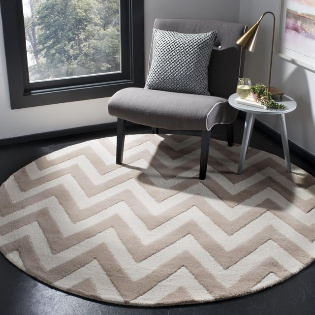 Safavieh Cambridge Kaitlyn Zig Zag Stripes Area Rug in a round shape with beige and cream zig zag stripes, placed under a gray upholstered chair with a patterned cushion, next to a small round side table with a brass lamp and a glass of white wine in a bright corner near a window, suitable for adding classic style and warmth to home decor.