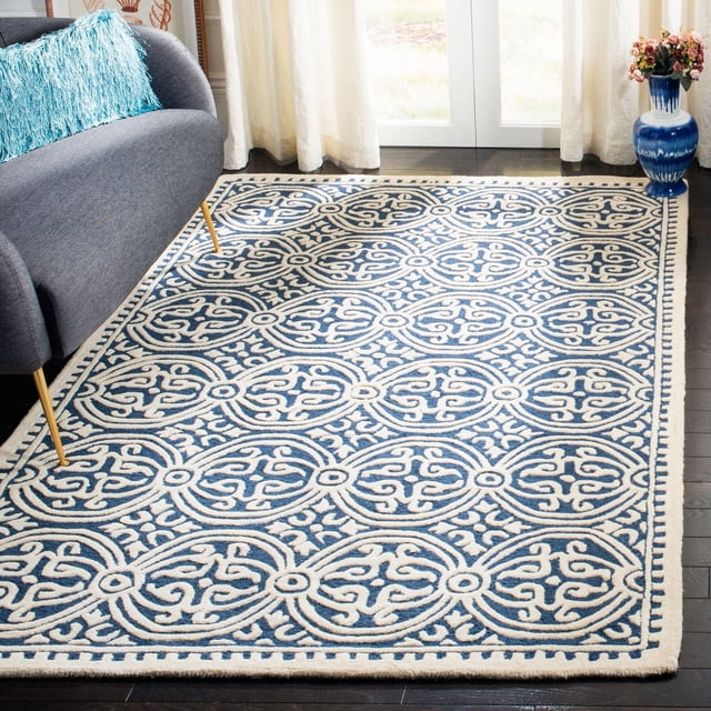 SAFAVIEH Cambridge Leslie Geometric Area Rug in blue and cream hand-tufted wool with a repeating geometric pattern of interlocking ovals and scroll motifs, placed on dark hardwood flooring beside a gray sofa with a blue textured pillow, near sheer white curtains and a blue floral vase, ideal for adding classic American style to home or office spaces.